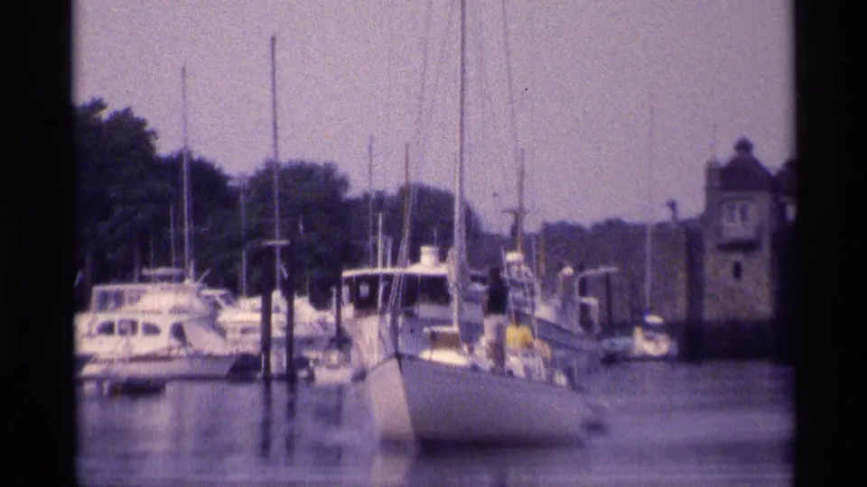 A closer 1973 New York marina frame shows sailboats, dock pilings, distant people on the dock, and a stone harbor structure.