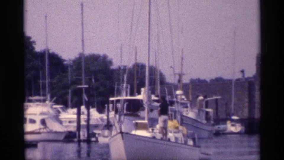 1973 New York 8mm frame showing sailboats, motor yachts, masts, water, and people aboard a white boat in a marina.