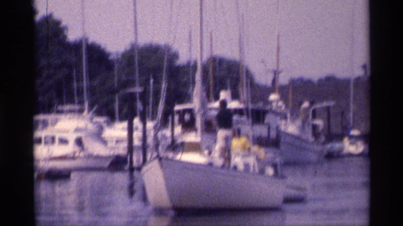 1973 New York 8mm frame of a white sailboat near docked yachts, with people visible on deck and a wooded shoreline behind.