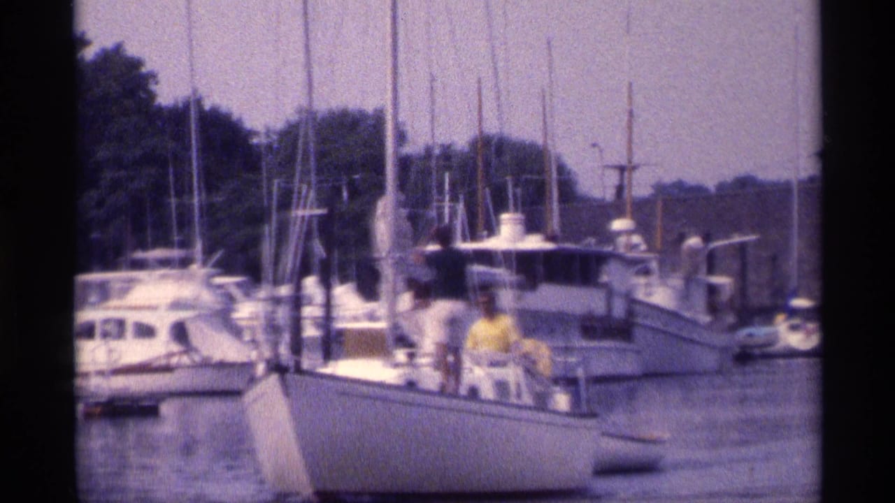 Soft 8mm frame from 1973 New York showing a marina, clustered masts, docked boats, and people on a white sailboat.