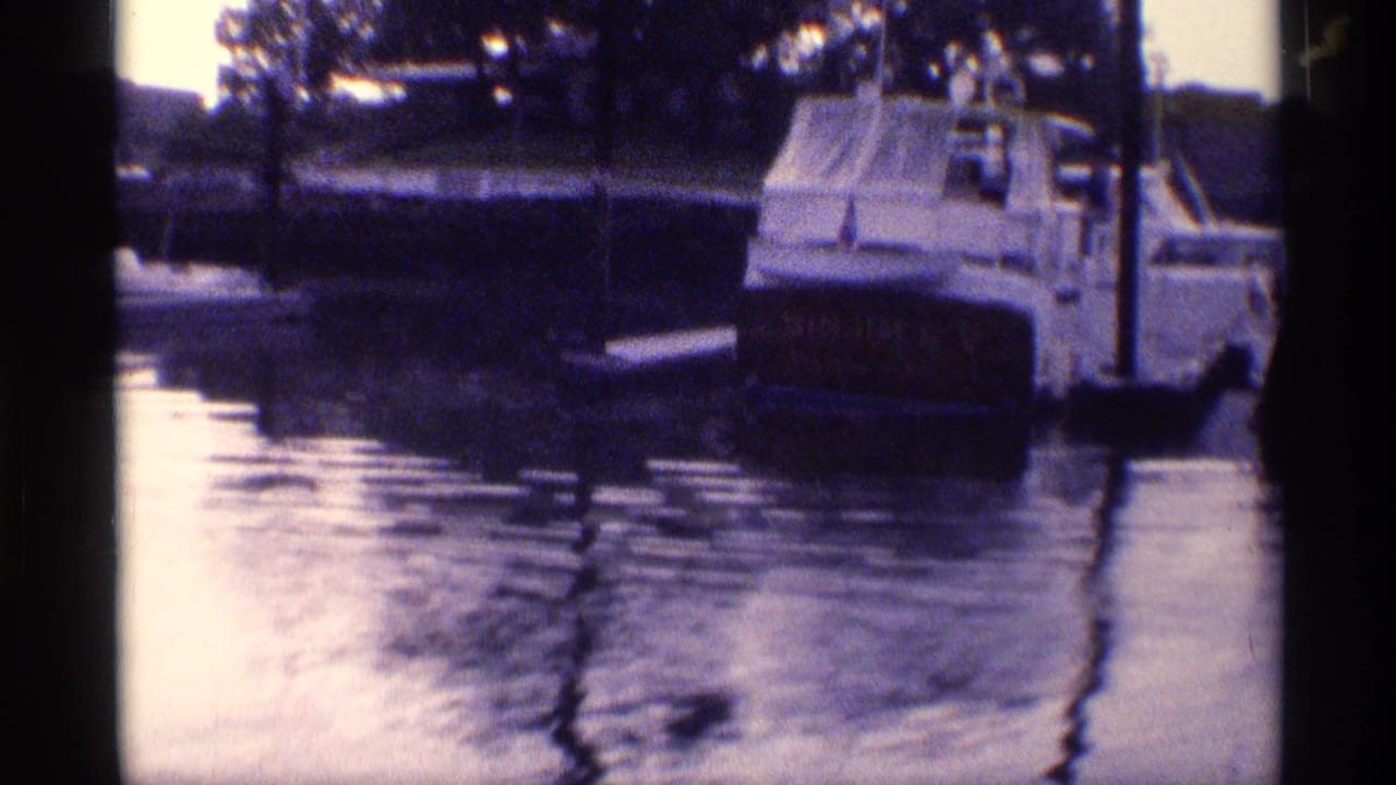 1973 New York 8mm frame showing a docked motor yacht, reflective water, posts, and trees along the marina edge.