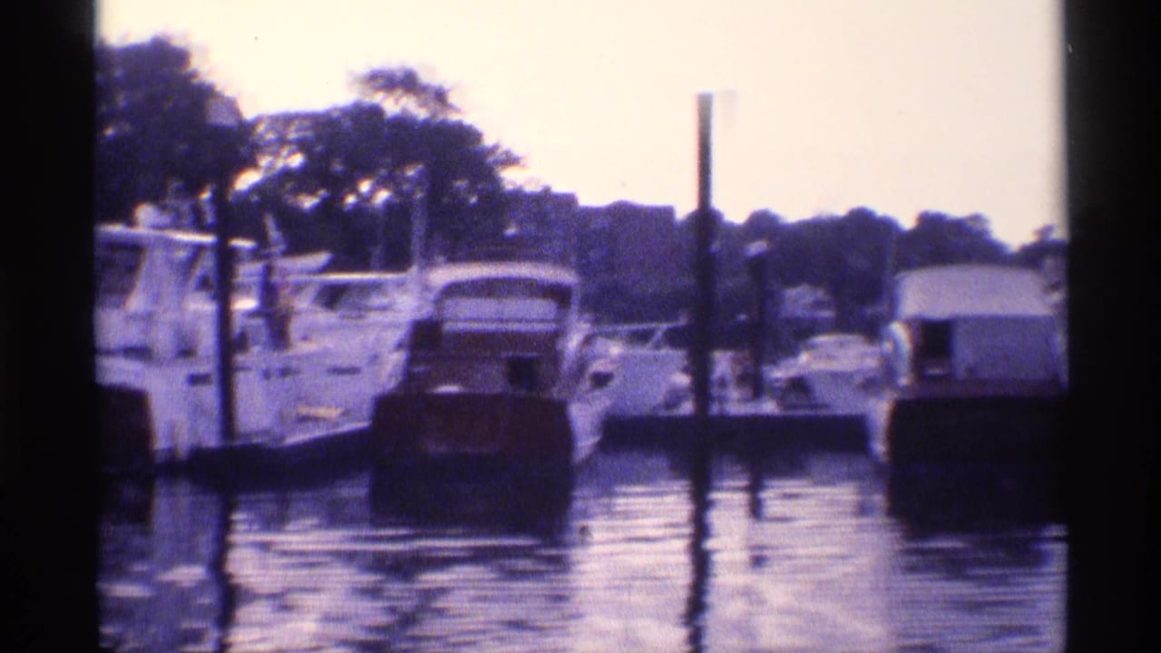 Docked motor yachts and small craft line a New York marina, with pilings, trees, and water reflections visible in the 1973 home-movie frame.