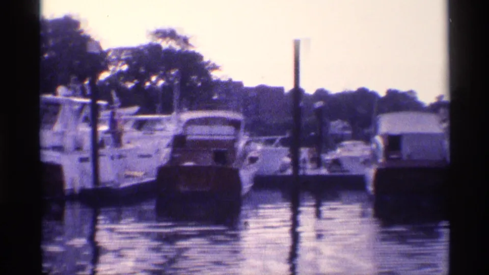 Docked motor yachts and small craft line a New York marina, with pilings, trees, and water reflections visible in the 1973 home-movie frame.