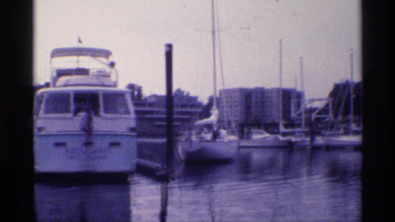 A white motor yacht is docked near sailboat masts and marina pilings in this 1973 New York 8mm waterfront view.