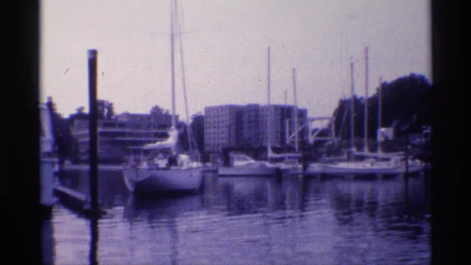 1973 New York 8mm frame showing moored sailboats, vertical masts, calm water, and apartment blocks beyond the marina.