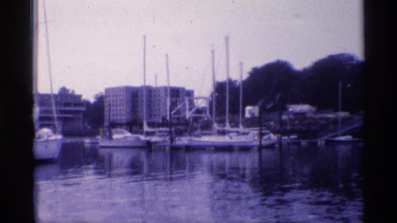 1973 New York waterfront frame with docked sailboats and their masts set against trees and mid-century apartment buildings.