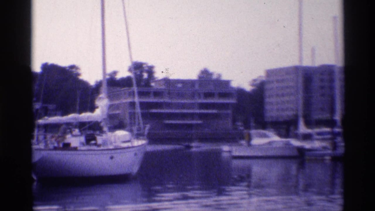 Closer 1973 New York 8mm view of a moored sailboat, calm reflective water, and apartment buildings along the waterfront.