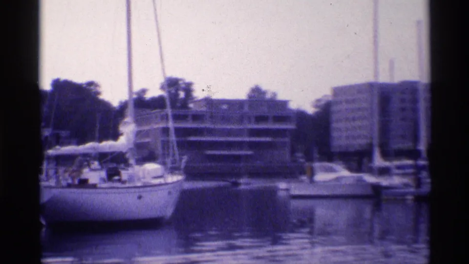 Closer 1973 New York 8mm view of a moored sailboat, calm reflective water, and apartment buildings along the waterfront.