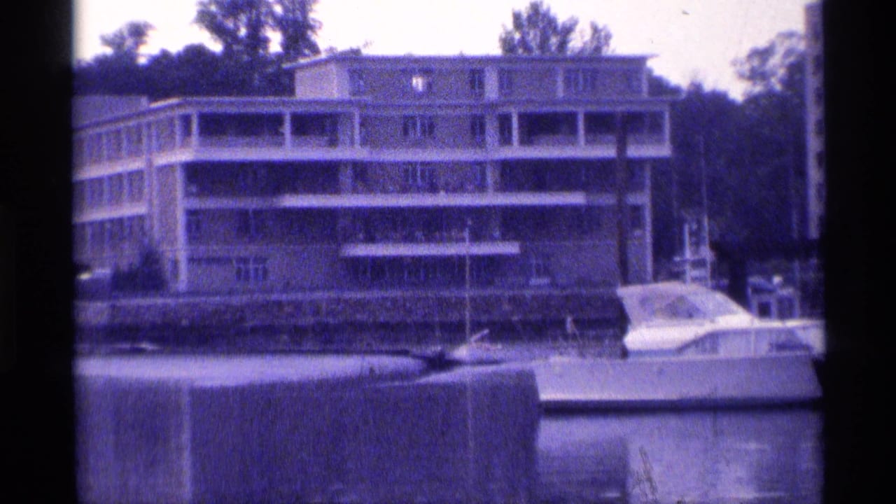 1973 New York marina frame showing a waterfront apartment building, moored motorboat, dock edge, trees, and still water.