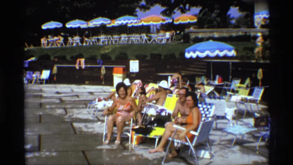 1973 New York poolside scene with adults lounging near folding chairs, towels, and striped umbrellas.