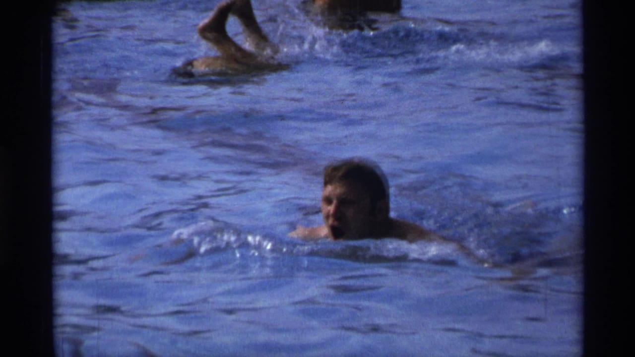 1973 New York pool footage frame with a man swimming toward the camera area while other bathers move through the water nearby.