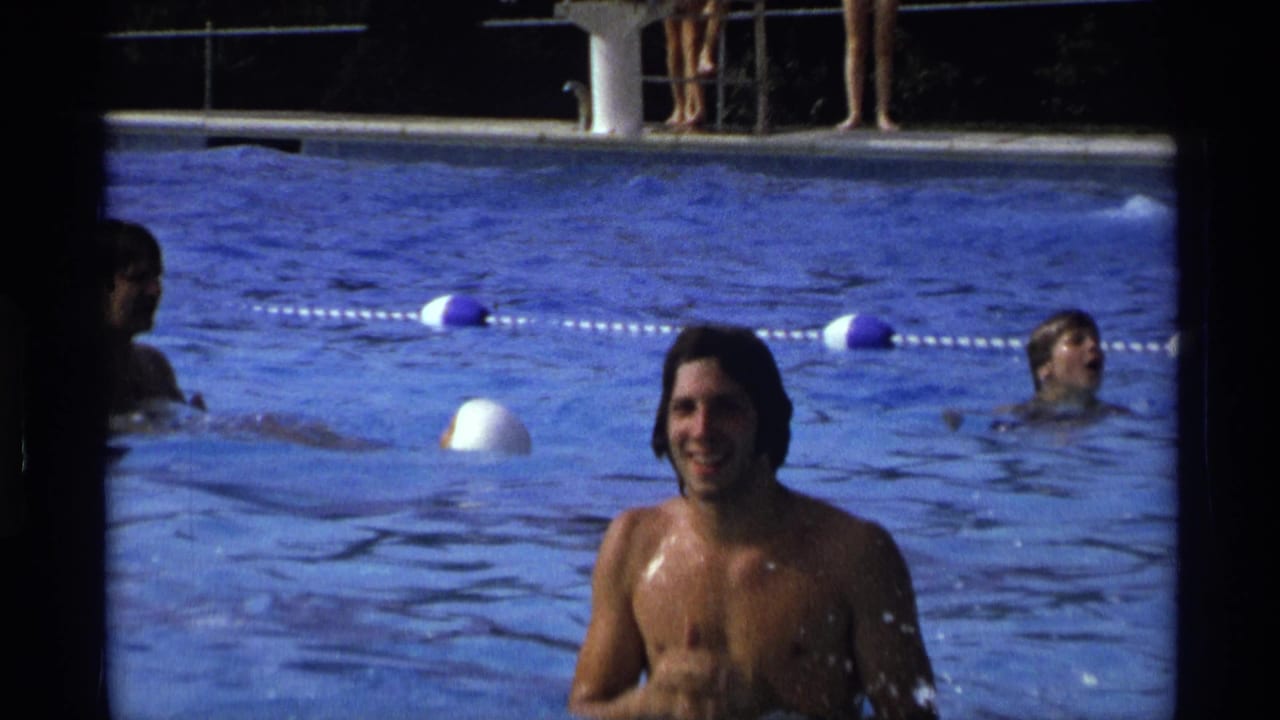 Young long-haired man smiling in a blue swimming pool with other swimmers behind him.