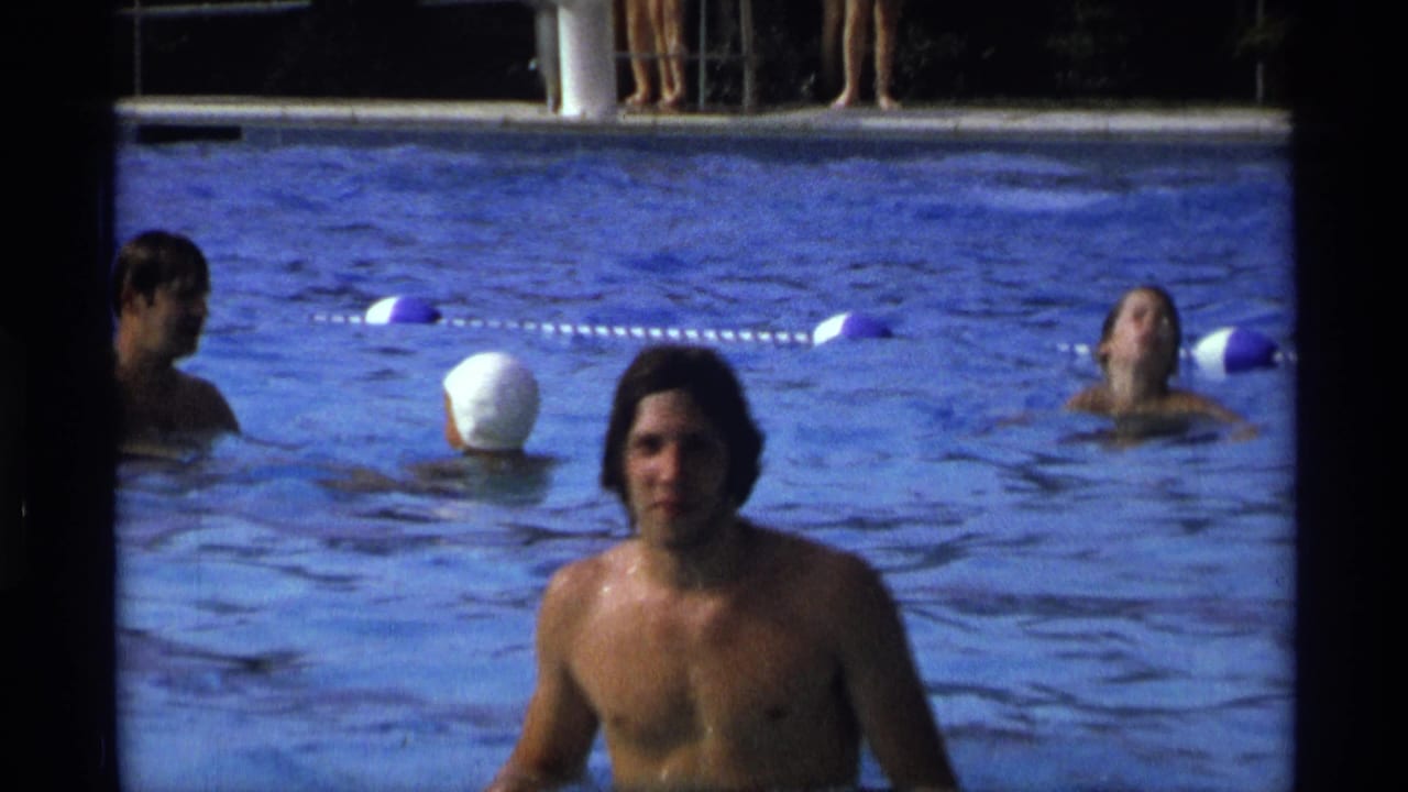 A young man faces the camera in a New York swimming pool, with blue and white lane floats and several swimmers visible in the 1973 home-movie frame.
