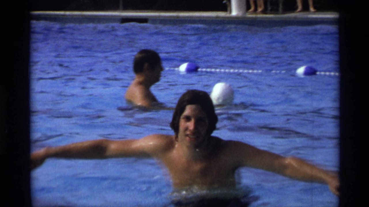 The young man holds the pool edge with both arms out in a 1973 New York pool scene, while another swimmer and floating lane markers appear behind him.