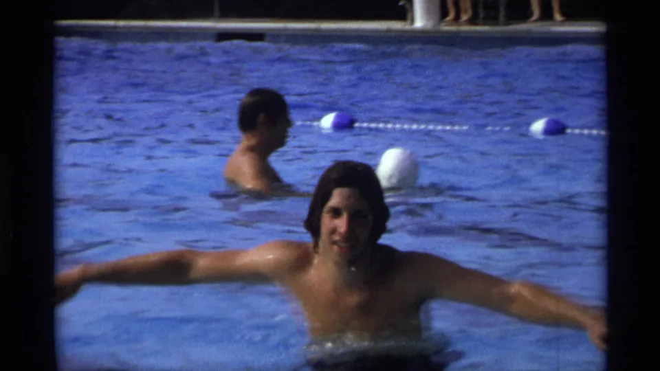 The young man holds the pool edge with both arms out in a 1973 New York pool scene, while another swimmer and floating lane markers appear behind him.