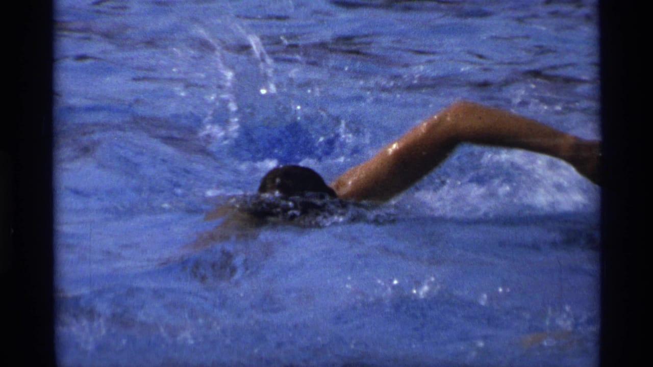 A swimmer moves through blue pool water in a front-crawl stroke, creating splashes in this short 1973 New York 8mm home-movie moment.