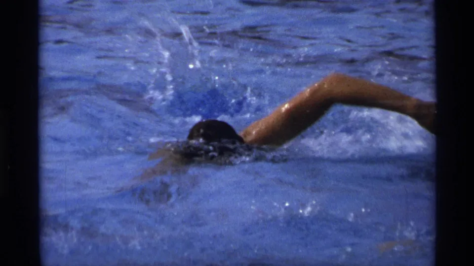 A swimmer moves through blue pool water in a front-crawl stroke, creating splashes in this short 1973 New York 8mm home-movie moment.