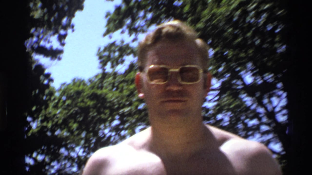 Shirtless man in gold-rimmed sunglasses faces the camera under trees.