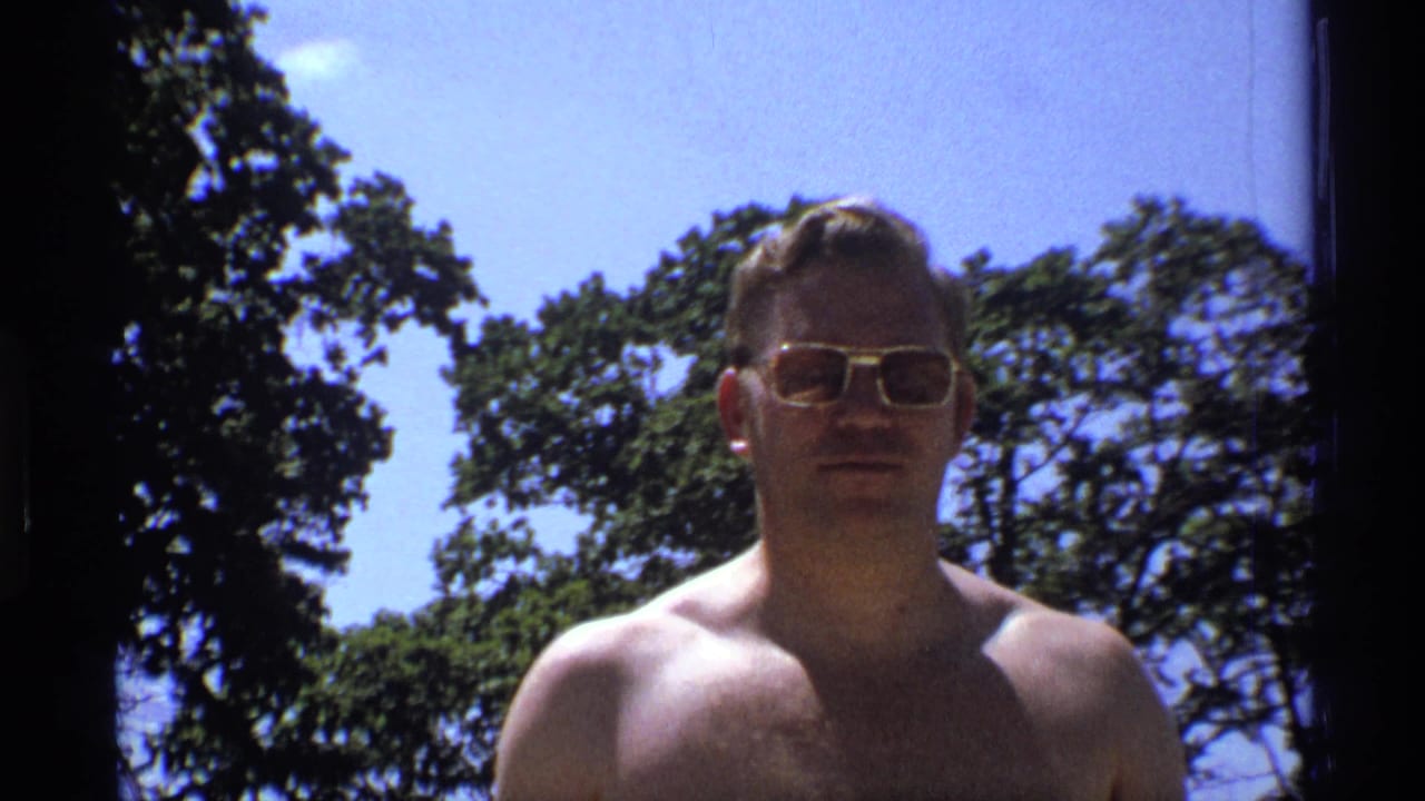 A shirtless adult man wearing gold-rimmed sunglasses stands outdoors in 1973 New York footage, with trees and clear sky behind him.