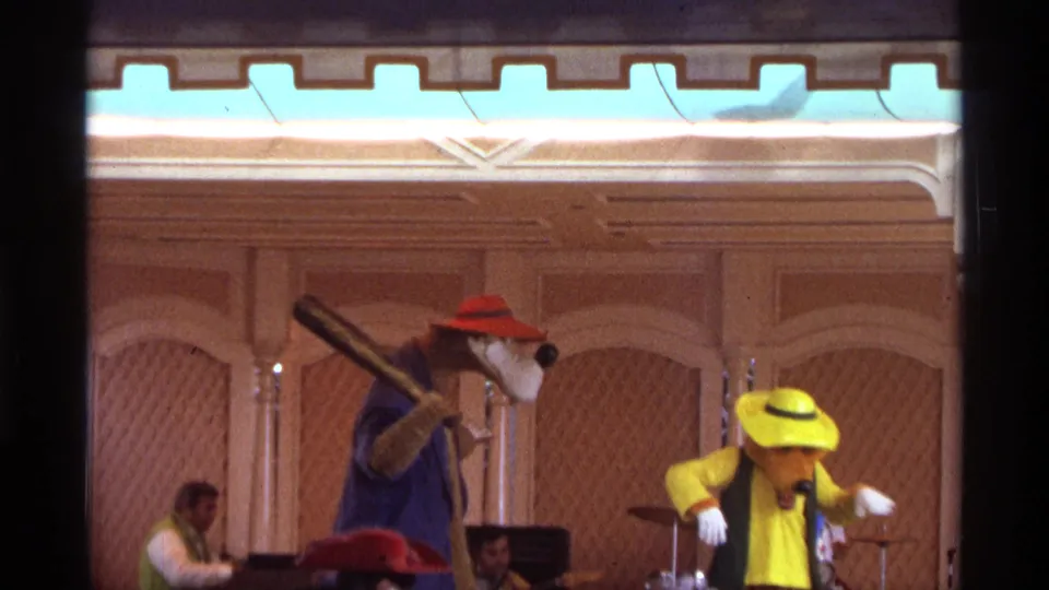 1974 Orlando home-movie frame of a costumed stage skit, with a bear character lifting a prop bat beside a yellow-hatted performer.