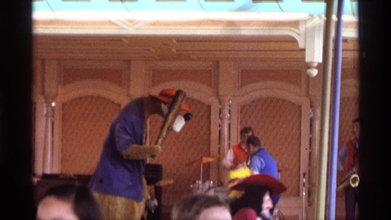 Costumed bear-like performer dances on a wood-paneled stage with musicians behind.