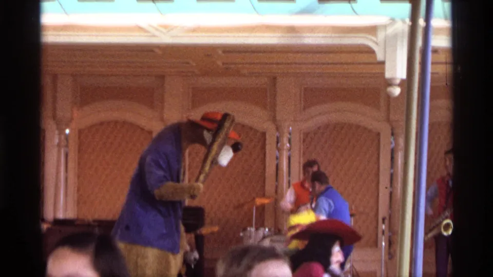 1974 Orlando 8mm frame of a costumed performer in a blue jacket and orange hat dancing before a live band on an indoor stage.