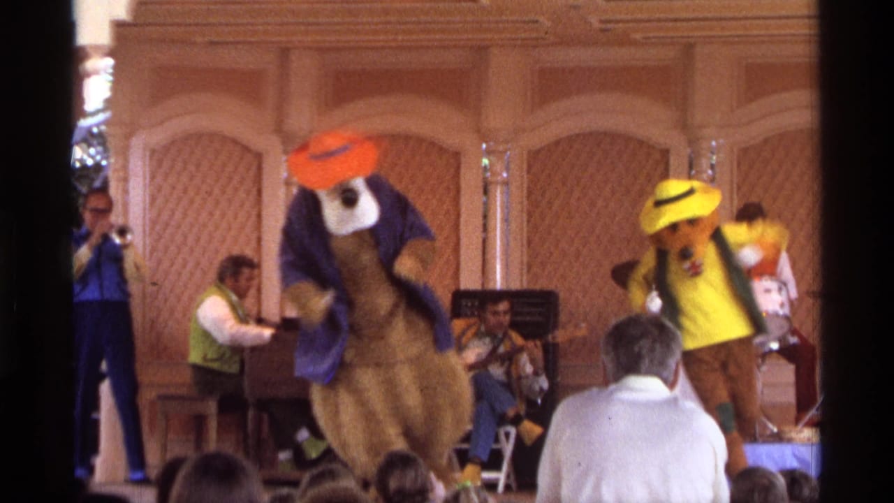 1974 Orlando stage-show frame with two costumed characters in bright hats dancing in front of trumpet, piano, and seated musicians.
