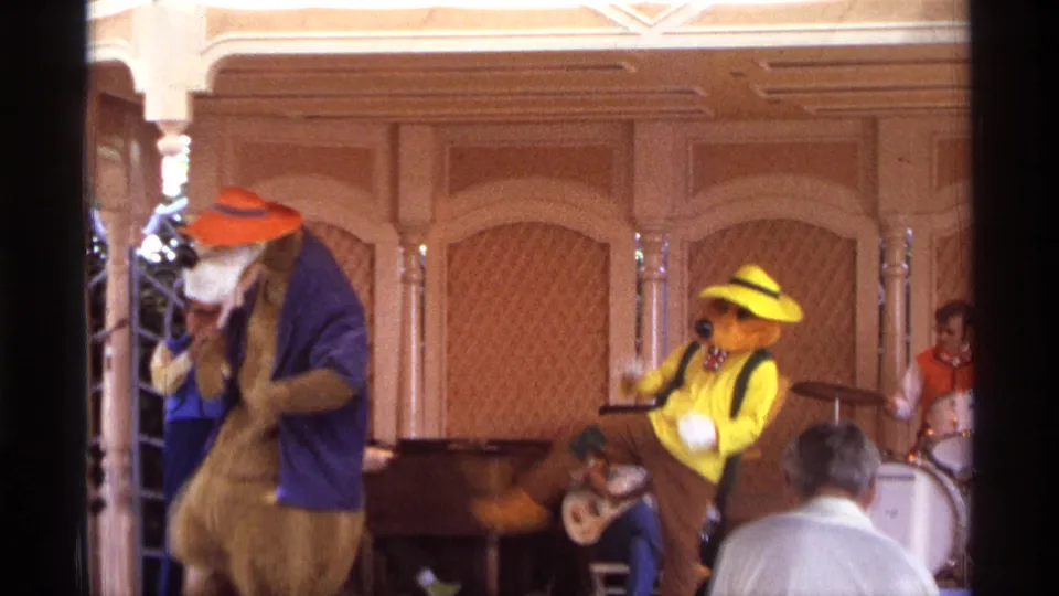 1974 Orlando 8mm frame showing two colorful costumed characters performing near a drum kit and guitar player on a wood-paneled stage.