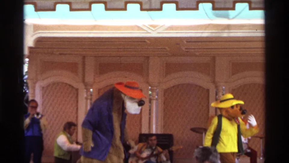1974 Orlando home-movie frame of costumed stage performers dancing with live musicians and audience heads visible in the foreground.