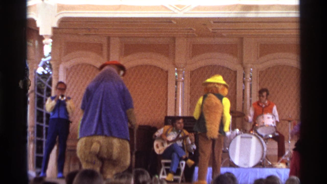 1974 Orlando frame with a large bear mascot turned away, a yellow-hatted animal performer, seated musicians, drummer, and audience foreground.