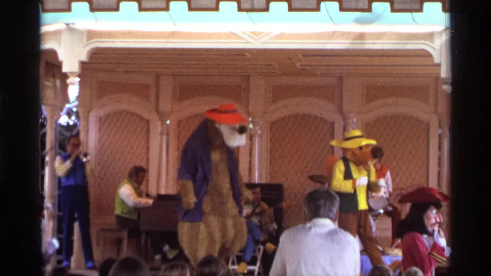 1974 Orlando home-movie frame showing a bear mascot and yellow-hatted performer on stage, with musicians and audience members visible in front.