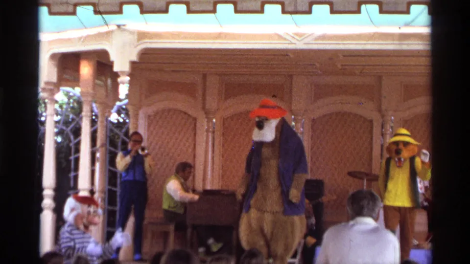 1974 Orlando frame with a bear mascot in a red hat near center stage, a yellow-hatted mascot at right, musicians at left, and audience heads below.