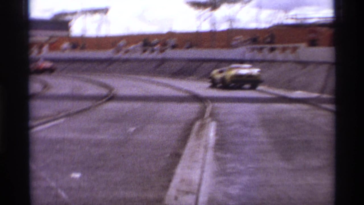 Yellow sports car enters a curved paved track with spectators behind a low wall.