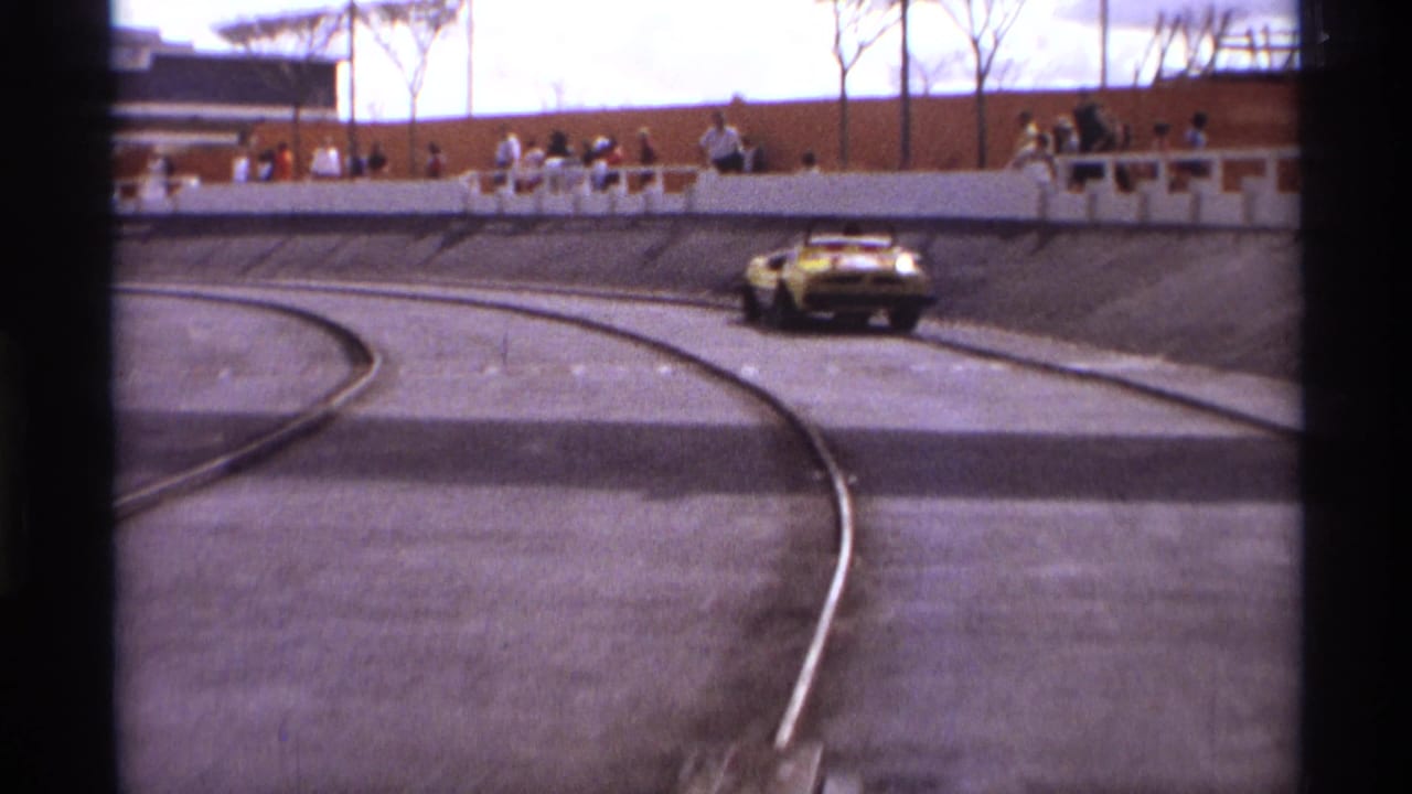 The yellow sports car rounds the paved curve in Orlando, Florida, with distant spectators lined along the wall in 1974 8mm footage.