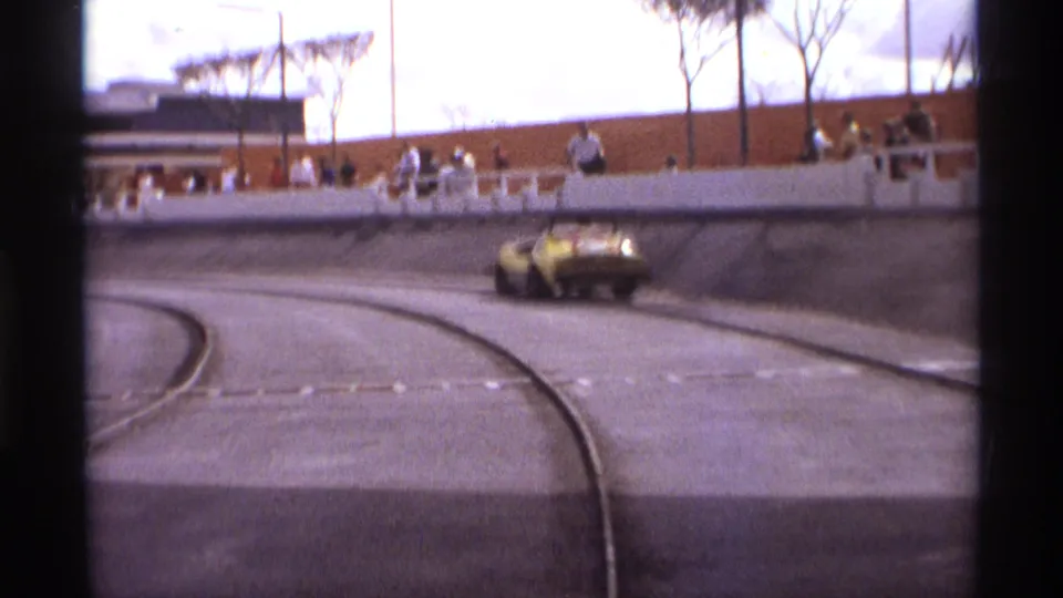 A yellow sports car passes through the turn on a paved track, with spectators standing behind the wall in 1974 Orlando footage.
