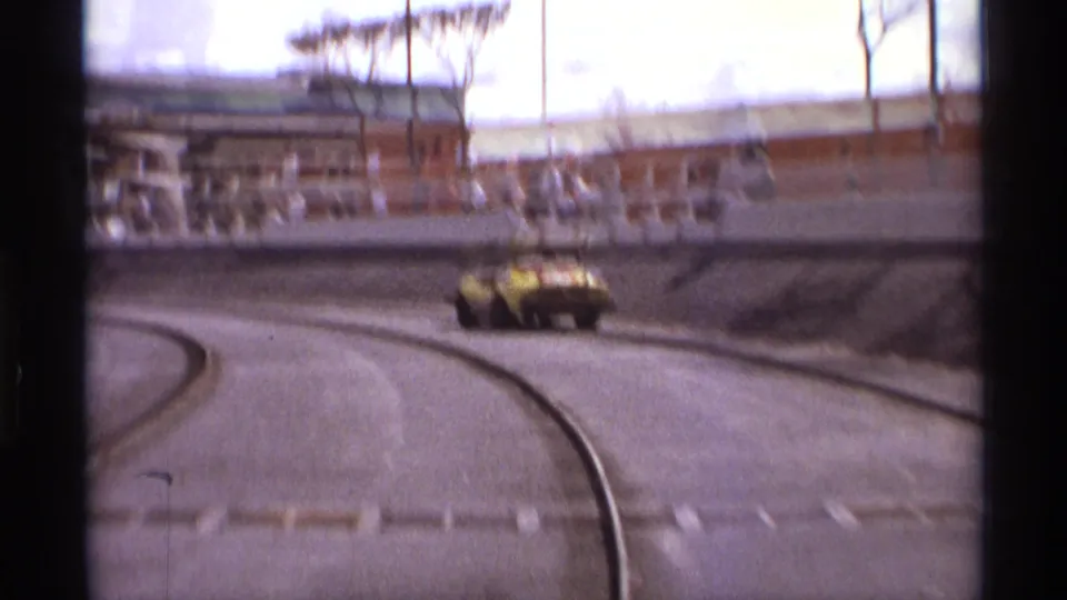 The yellow sports car continues out of the bend on the Orlando track, seen through faded 8mm color with spectators in the background.