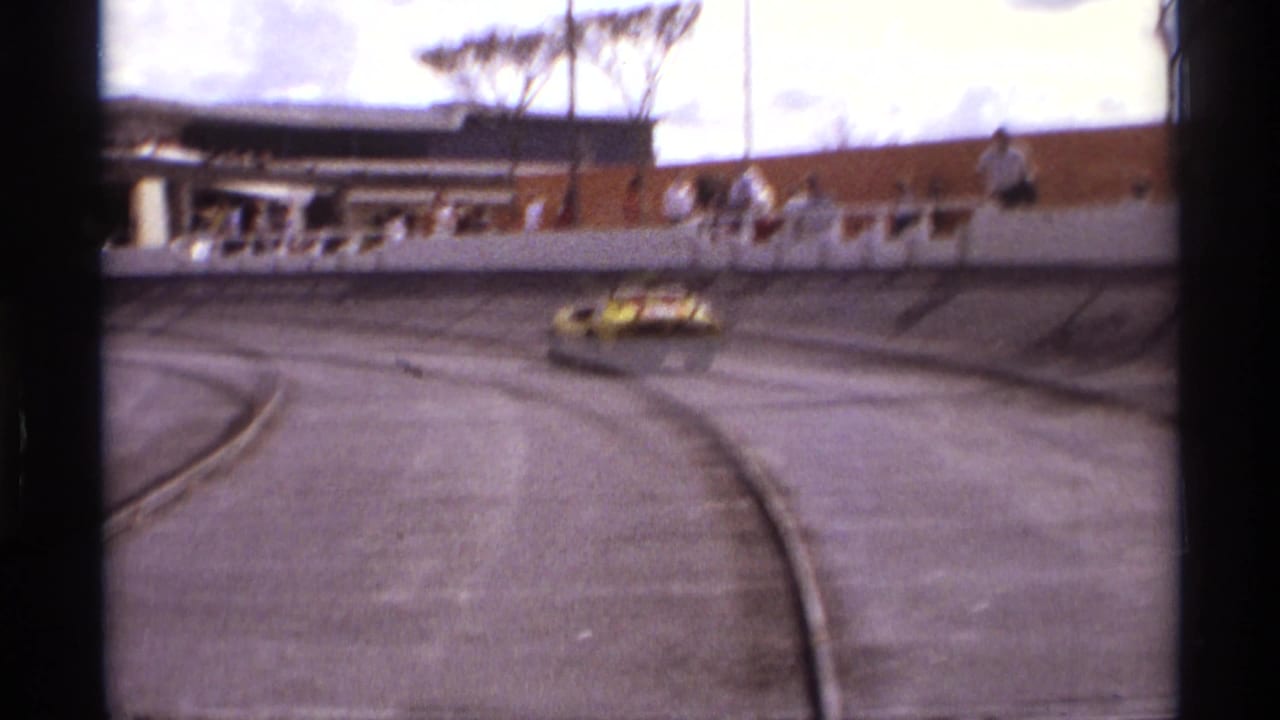 1974 Orlando 8mm frame showing a yellow race car entering a broad speedway turn with distant spectators above the track wall.