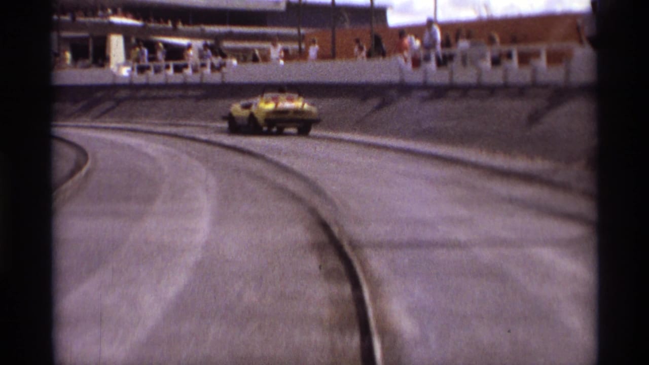 The yellow car appears near the outer edge of the Orlando speedway curve, with railings and spectators visible in the background.