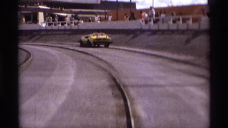 The yellow car appears near the outer edge of the Orlando speedway curve, with railings and spectators visible in the background.