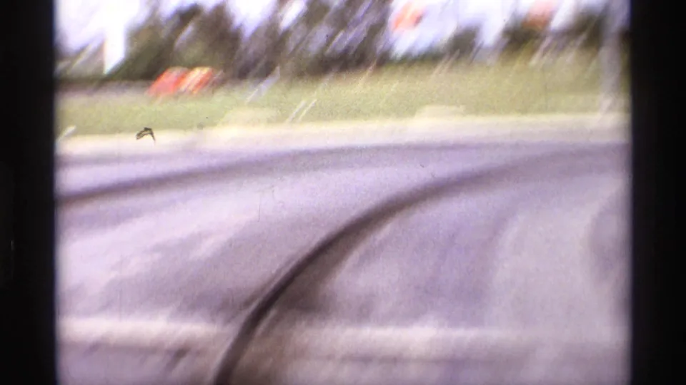 A motion-blurred 8mm frame shows the speedway curve, tire marks, grass beyond the pavement, and small flags or signs in the distance.