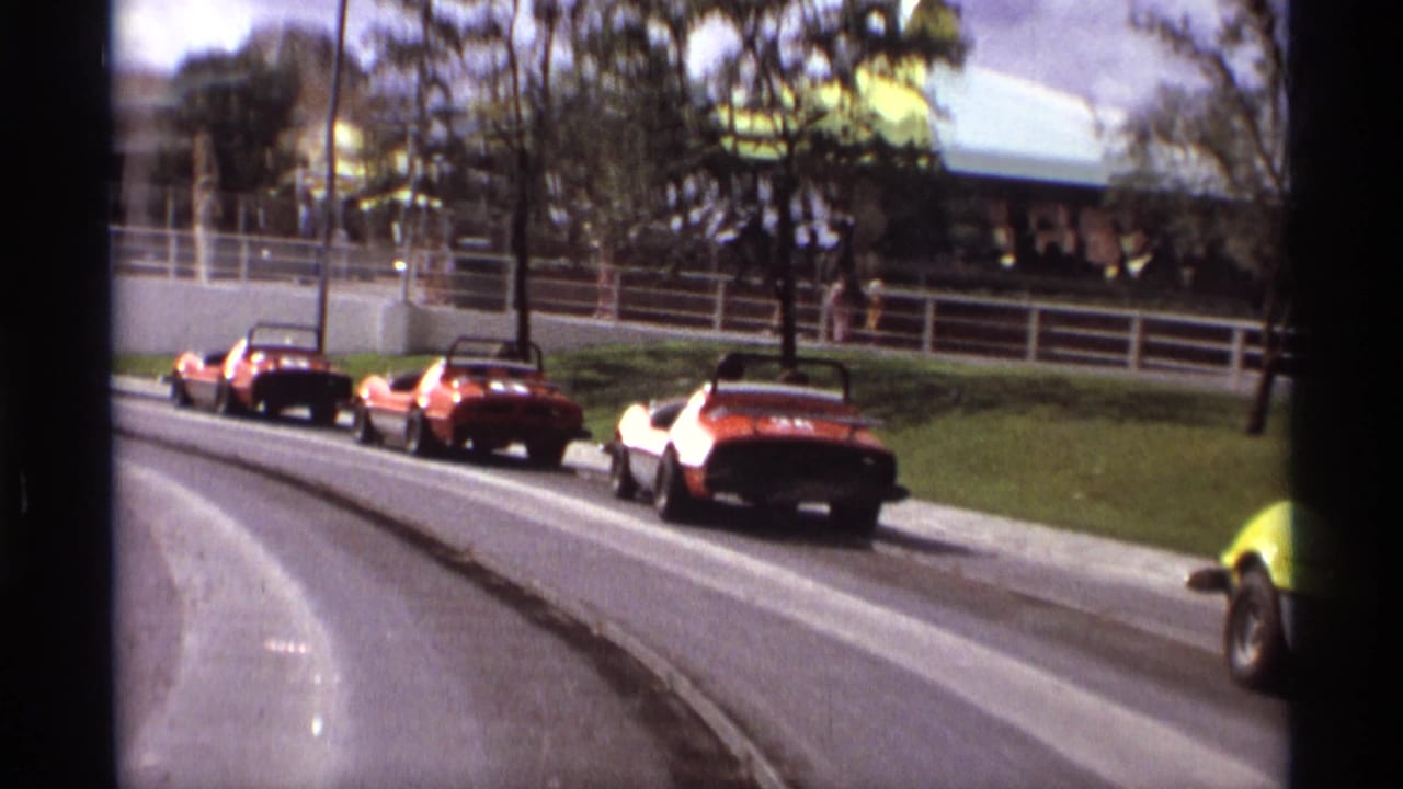 1974 Orlando amusement-park go-karts, mostly red and orange, move through a curve with railings, trees, and park structures behind them.