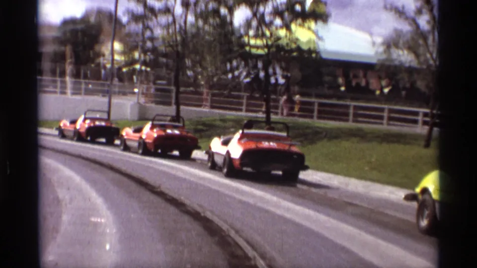 1974 Orlando amusement-park go-karts, mostly red and orange, move through a curve with railings, trees, and park structures behind them.