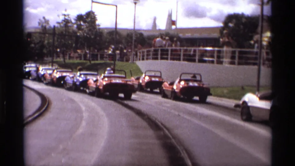 1974 Orlando frame with a longer line of go-karts on a curving paved track, bordered by grass, white railings, trees, and park structures.