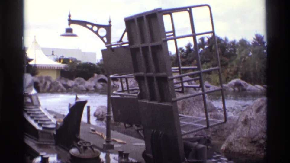 1974 Orlando 8mm frame showing a dark themed ride vehicle near water, artificial rocks, palm trees, and a white tent-like structure.