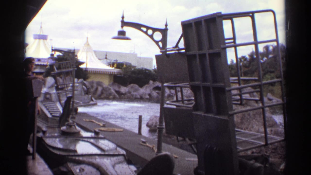 Grey ride boat structure beside a waterway with rocks, lamppost, and white tented roof in the background.