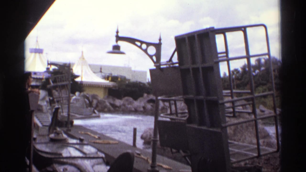 1974 Orlando home-movie frame looking from a moving ride toward a grey barge-like structure, water, rocks, lamp, and white tented roof.