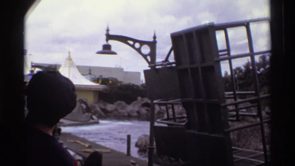 1974 Orlando ride POV with a dark partial rider silhouette, grey boat structure, rocky waterway, ornate lamp, and distant tented roof.