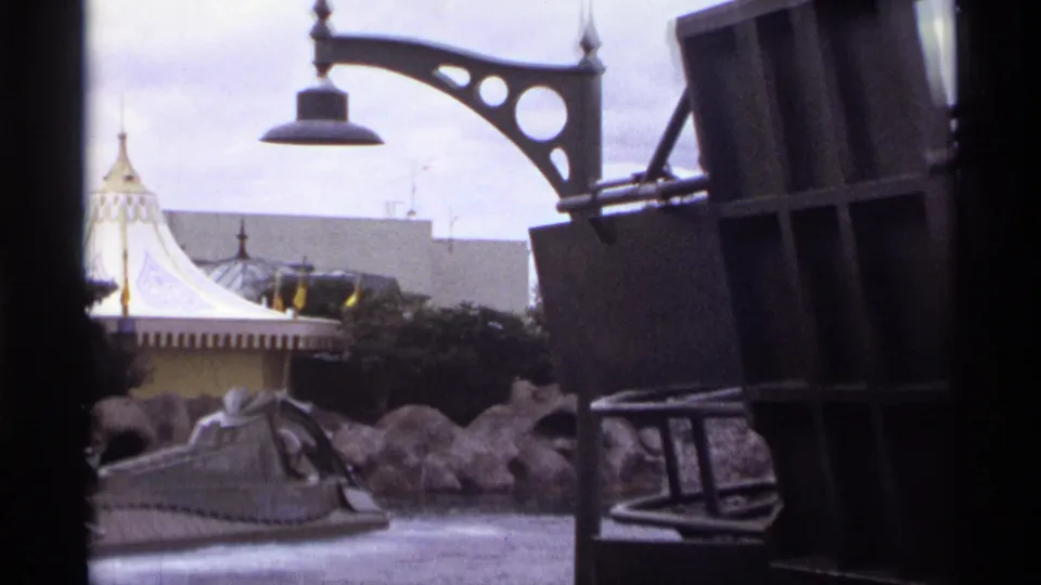 1974 Orlando 8mm ride frame with a close decorative lamppost, grey structure, rocky waterway, and white tented building beyond.