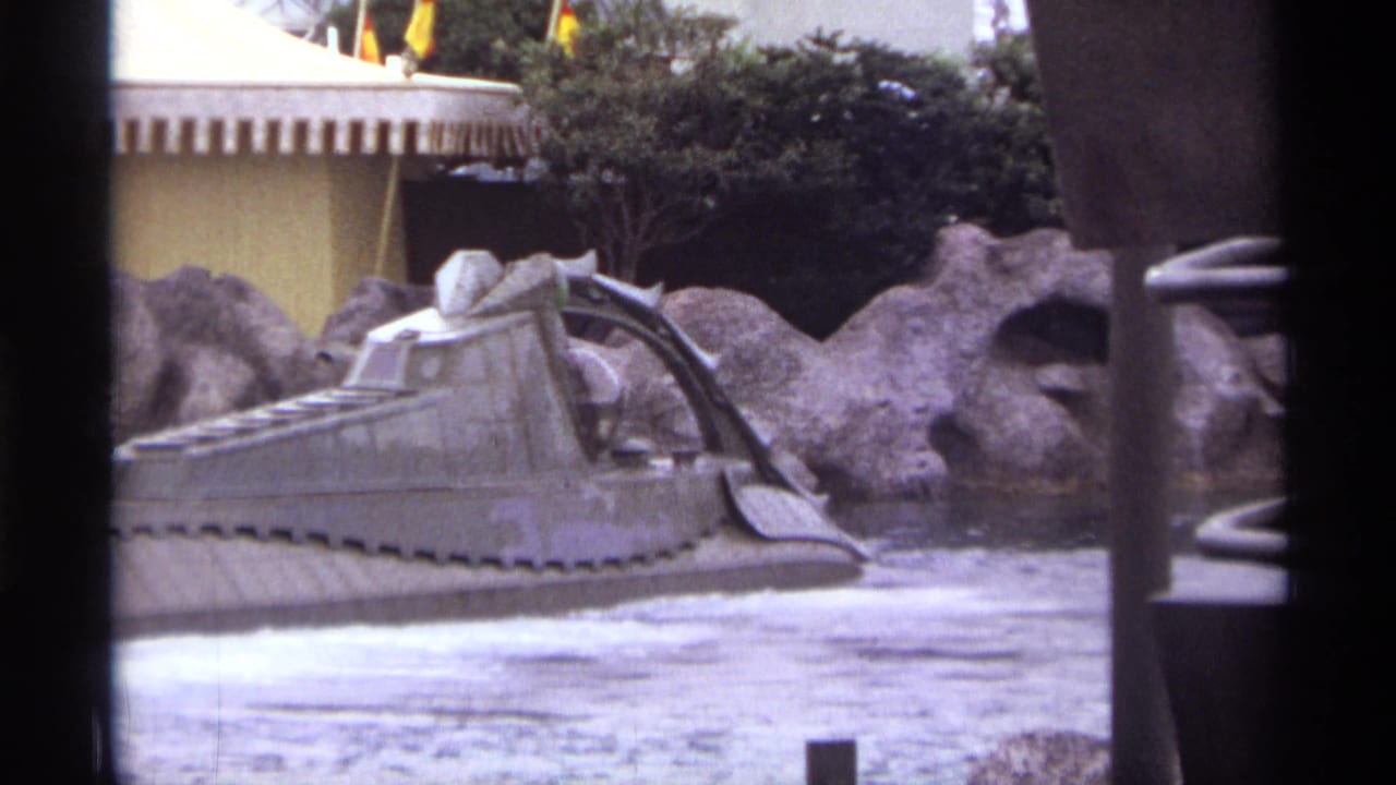 Gray tracked water-ride vehicle beside artificial rocks and foamy water.