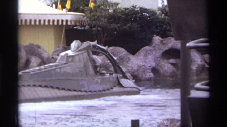 1974 Orlando 8mm frame of a gray tracked amphibious-style vehicle moving along a waterway near artificial rockwork.